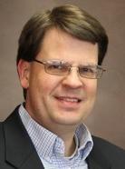 A man with glasses, short brown hair, and wearing a suit jacket and checkered shirt, smiling against a plain brown background.